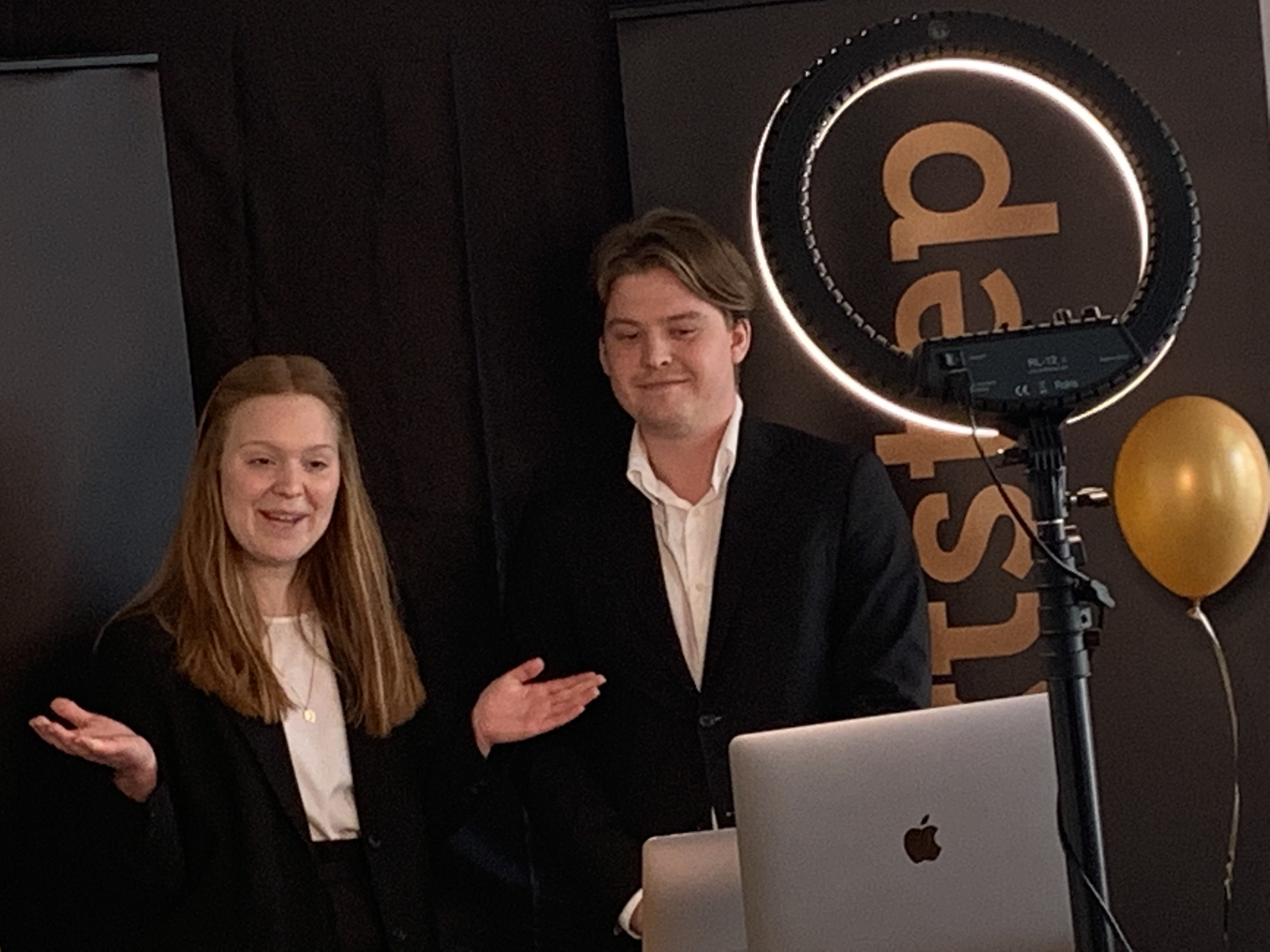Two students stand infront of a black backdrop presenting towards a laptop. A ringlight can be seen in the foreground.