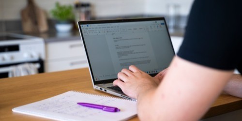 A person writing with a purple pen in a notebook next tto a laptop