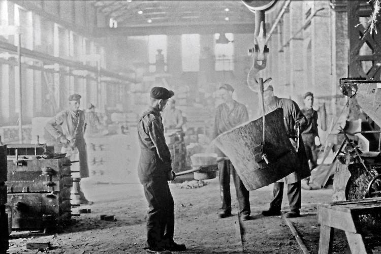 Svartvit bild där en fabriksarbetare står vid ett stort rykande kärl i JMW-hallen. Runt om står fler fabriksarbetare.