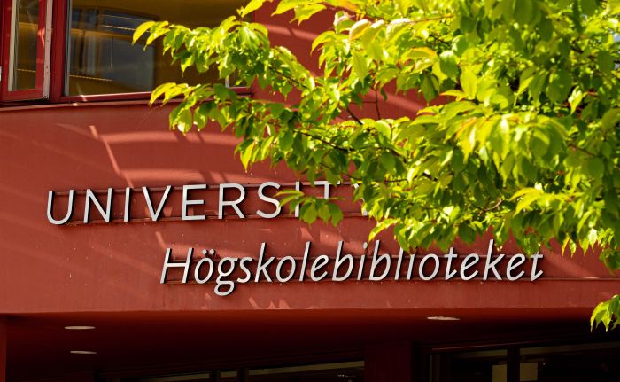 Gröna löv framför en röd husfasad där det står Högskolebiblioteket.